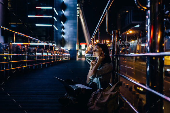 side view of woman listening music in headphones with laptop on knees on city street at night - Stock Photo - Images