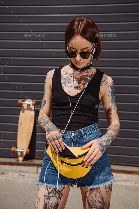 stylish tattooed woman listening music in earphones and putting smartphone in waist bag