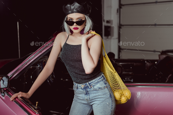 stylish blonde girl in sunglasses and beret holding string bag with lemons and looking at camera
