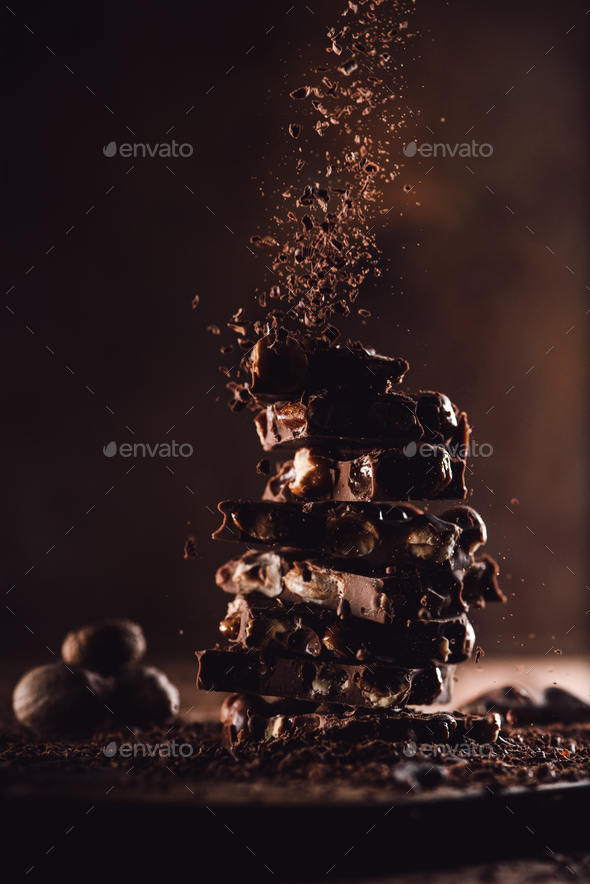 close up shot of nutmegs and grated chocolate falling on stack of chocolate pieces on wooden table