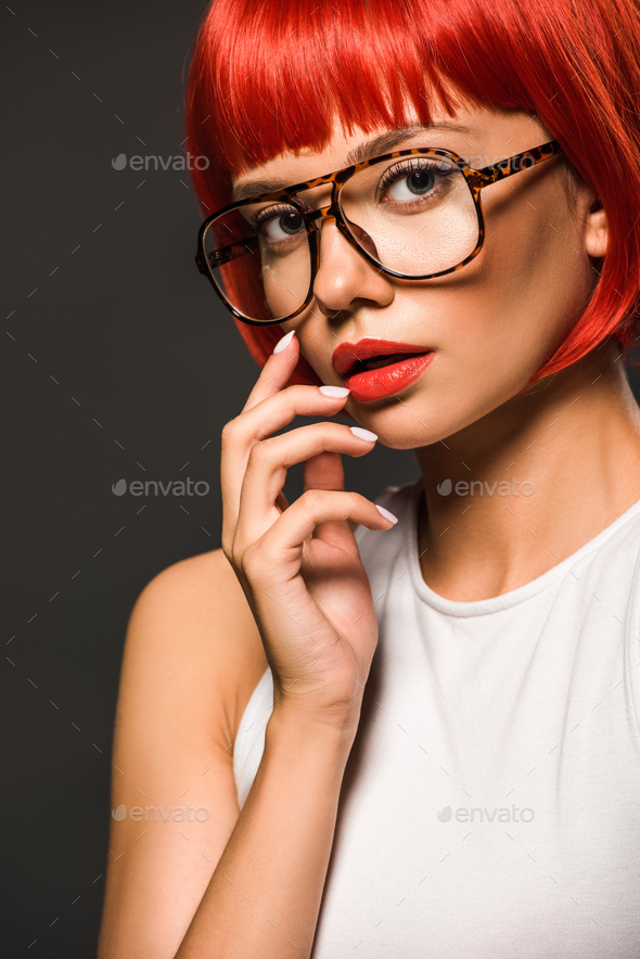 beautiful young woman with red bob cut and stylish eyeglasses looking at camera isolated on grey
