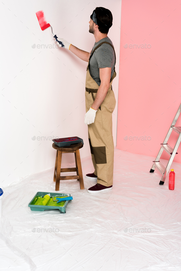 young man in working overall and headband painting wall in red by paint roller near chair with