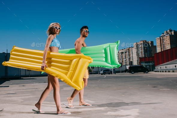 attractive young couple in beach clothes with inflatable beds walking on parking
