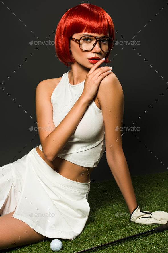 seductive young woman with red bob cut sitting on green grass with golf equipment and looking at