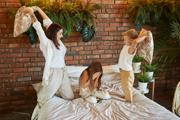 Family leisure. Mom son and daughter fight on pillows on the bed in the bedroom. Joy and fun