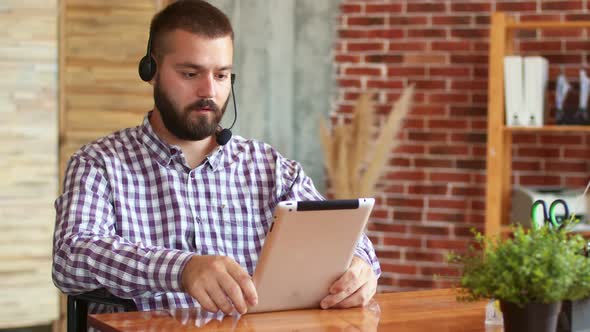 Hipster Man Wearing Headphones with Microphone is Sitting in Office and Using Electronic Tablet Side alt