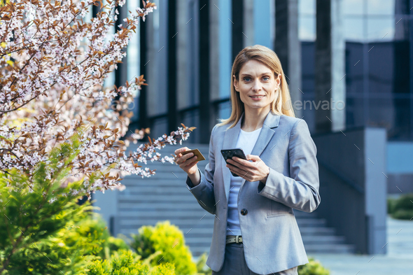 Happy business woman near office building looks at camera and smiles, uses phone