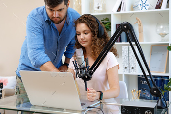 Female radio host recording podcast in studio, man colleague helps her to adjust sound.