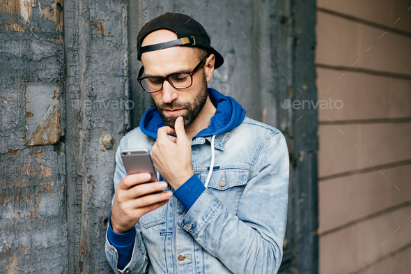 Serious concentrated bearded man in cap and denim jacket standing ...