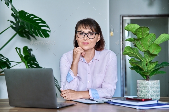Middle aged woman looking at camera sitting at her desk with laptop - Stock Photo - Images