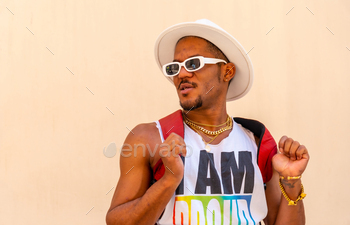 Portrait of a gay black ethnic man at the pride party on a cream wall, LGBT flag