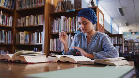 Asian female student wearing a blue hijab sitting at a desk with open books and taking notes alt