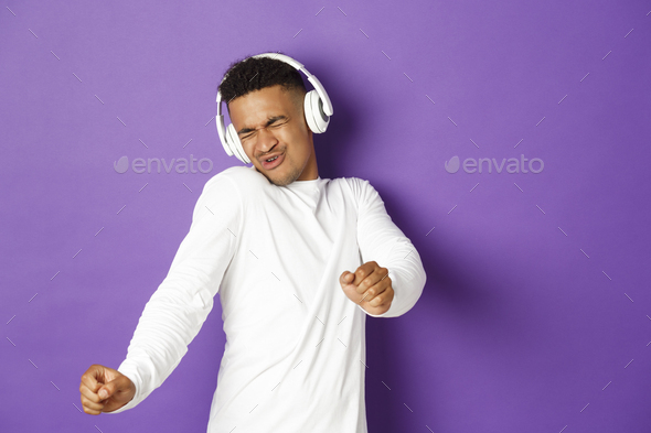 Image of carefree african-american guy dancing, listening music in wireless headphones, standing