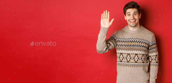 Portrait of handsome and friendly guy in christmas sweater, saying hello and waving hand, greeting