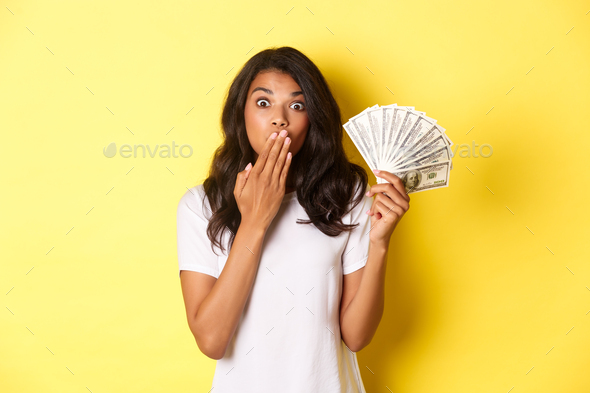 Image of surprised african-american girl winning money, gasping amazed, standing over yellow