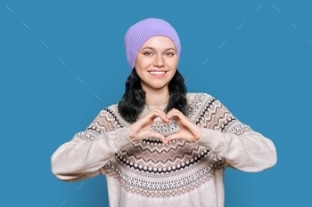 Young teenage female in hat sweater showing heart gesture with hands on blue background