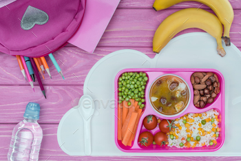top view of tray with kids lunch for school, pink bag, pencils and bottle of water on pink table