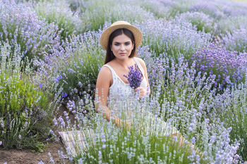 Beautiful woman in white dress sitting in violet lavender field and holding small bouquet