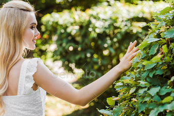 back view of beautiful smiling blonde woman touching green leaves