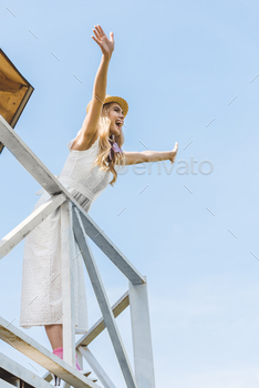 low angle view of cheerful young woman in dress and wicker hat raising hands and looking away