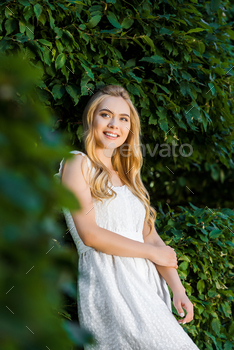 selective focus of beautiful smiling blonde girl in white dress posing near green leaves