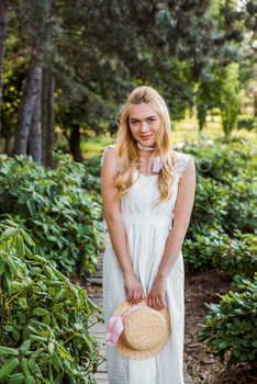 beautiful young blonde woman in white dress holding wicker hat and smiling at camera in park