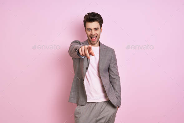 Happy young man in suit pointing camera while standing against pink background
