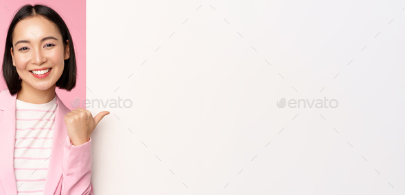 Portrait of smiling asian businesswoman in suit, corporate lady pointing finger at white empty wall