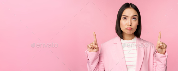 Upset asian corporate woman in office suit, pointing and looking up with disappointed face