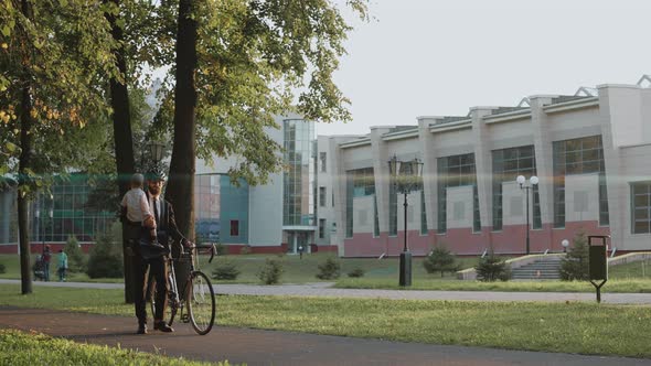 Businessman Walks with His Baby Sitting in Arms and Pushing Bike After Work Day alt
