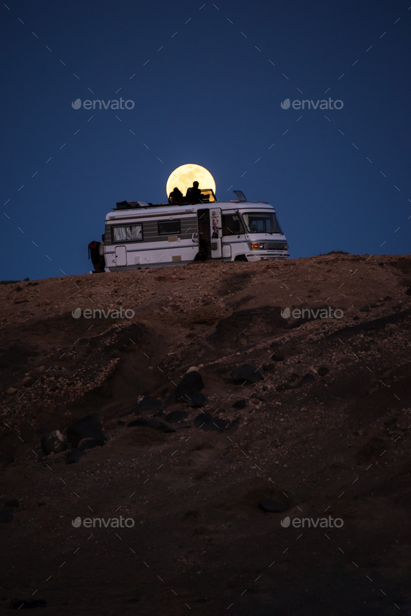 Silhouette of Couple on the Roof of a Motorhome in a Cliff with the Full Moon Behind.Vertical Image