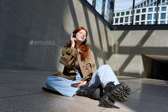 Teen redhead girl in headphones listening music using streaming app outdoor.