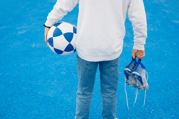 Child soccer player back with ball in his arm and boots in his hand