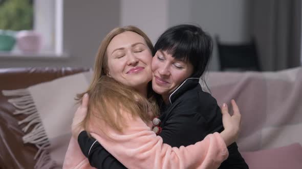 Two Happy Women Hugging Smiling Looking at Camera Sitting in Comfortable Living Room on Cozy Couch alt