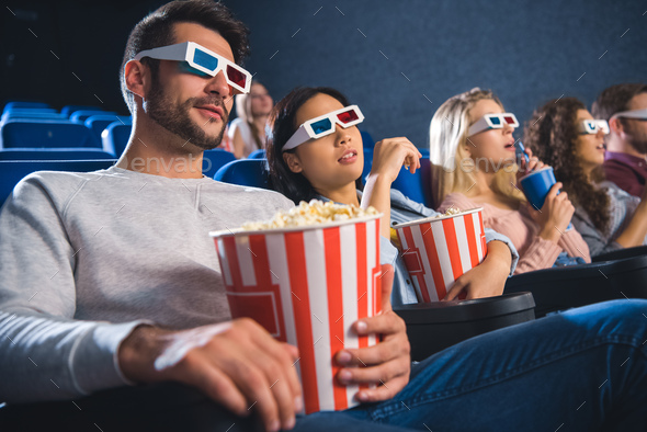 multiethnic friends in 3d glasses with popcorn watching film together in movie theater