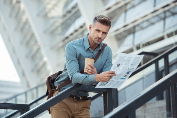 handsome middle aged man holding coffee to go and newspaper while standing on stairs