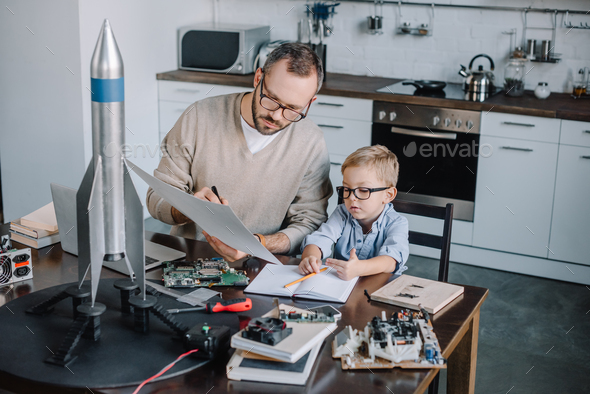 father and son modeling rocket and looking at blueprint at table in kitchen