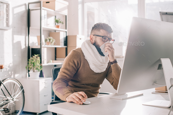 sick businessman in knitted scarf sitting in office, sneezing and covering mouth with tissue