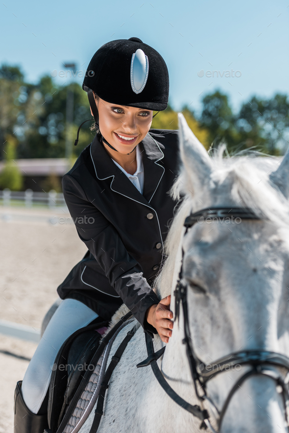 smiling attractive female equestrian in professional apparel riding horse at horse club
