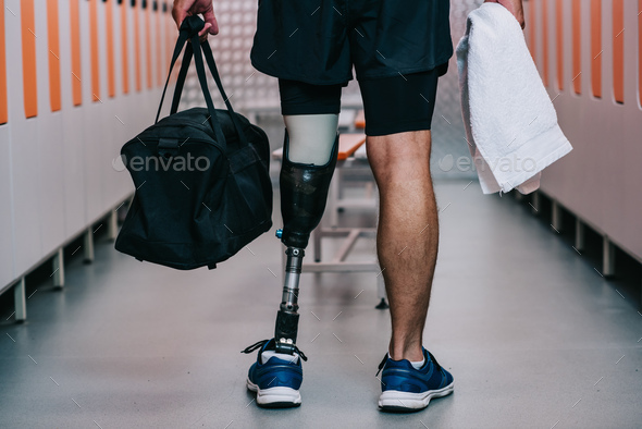 cropped shot of sportsman with artificial leg standing at gym changing room with towel and bag