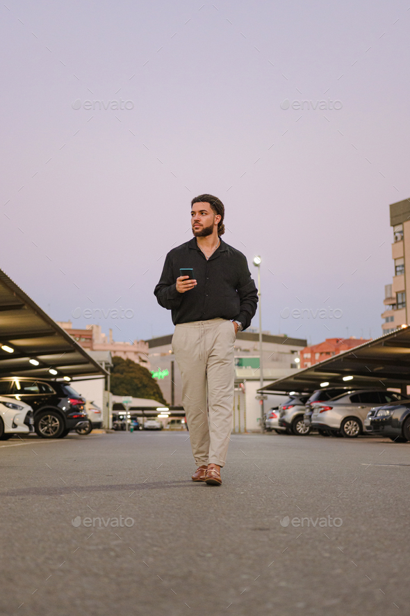 Latin man walks with a mobile phone in a parking lot at dusk