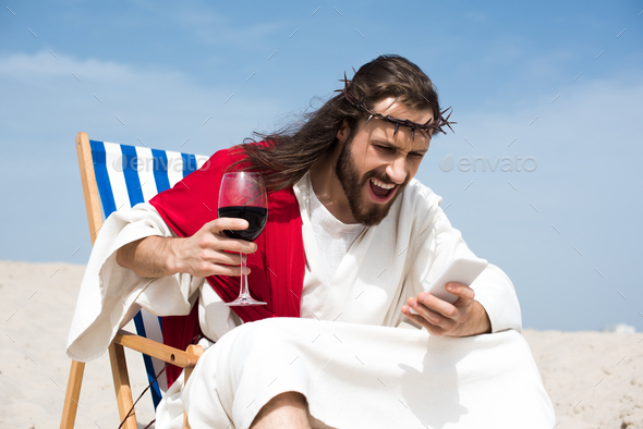 angry Jesus resting on sun lounger with glass of wine and screaming at smartphone in desert