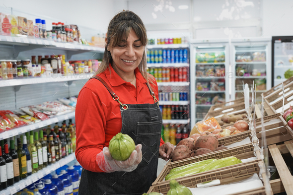 Mature latin woman working inside supermarket while holding fresh oragnic zucchini