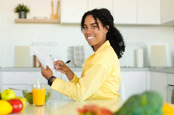 Diet plan concept. Happy black lady writing daily ration diet or menu in notebook, sitting at
