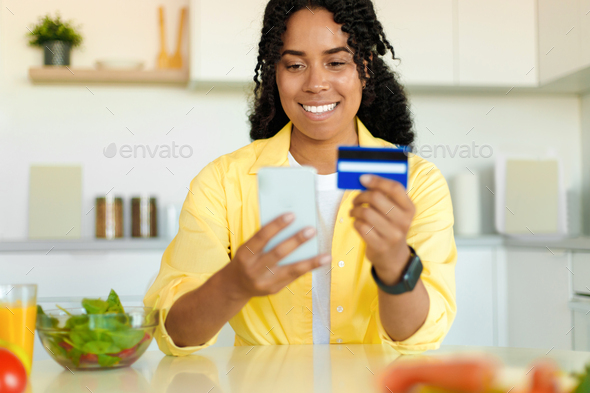 Cheerful black woman buying food online, using cellphone and credit card, purchasing ingredients for