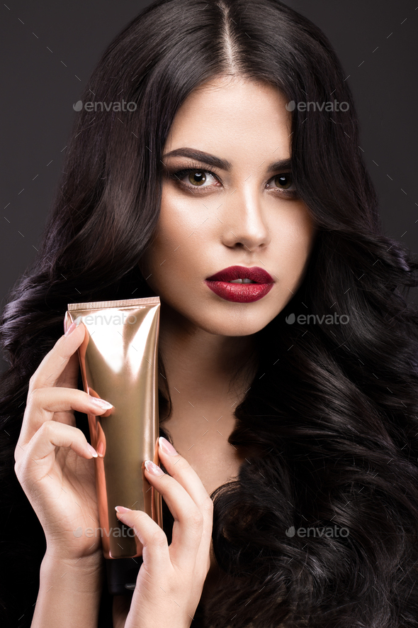 Beautiful brunette model: curls, classic makeup and red lips with a bottle of hair products.