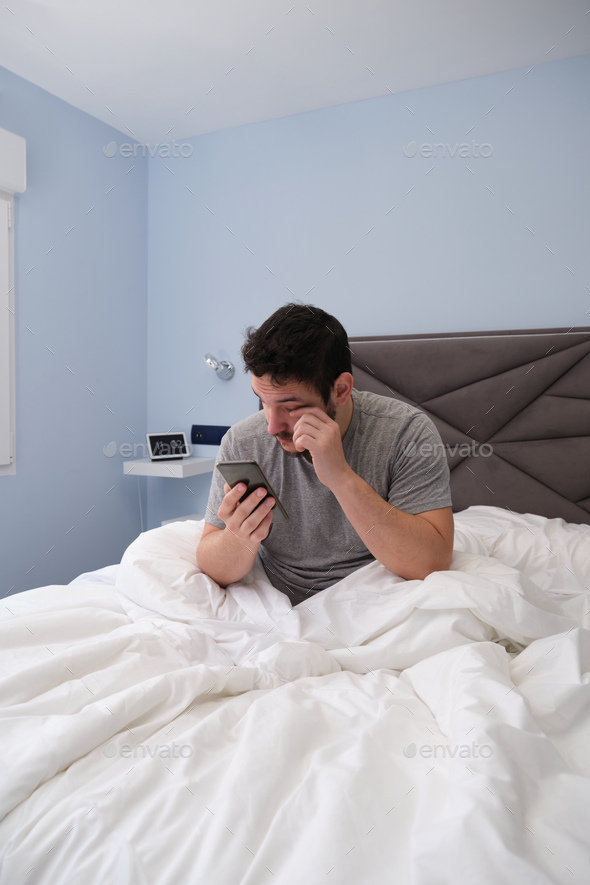 Young latin man rubbing his eyes in bed after sleeping and checking his smartphone