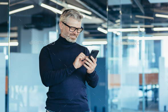 Senior gray-haired businessman in office using phone, happy investor reading message