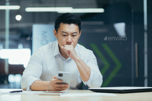 Confused Asian guy holding smart phone feels concerned thinking over received message. Mobile phone