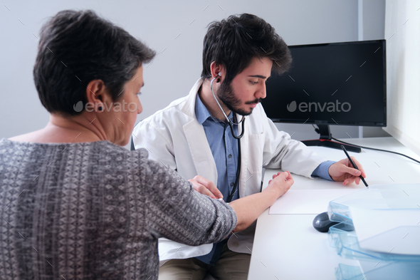Young doctor listening to mature woman patient heart with a stethoscope and writing down